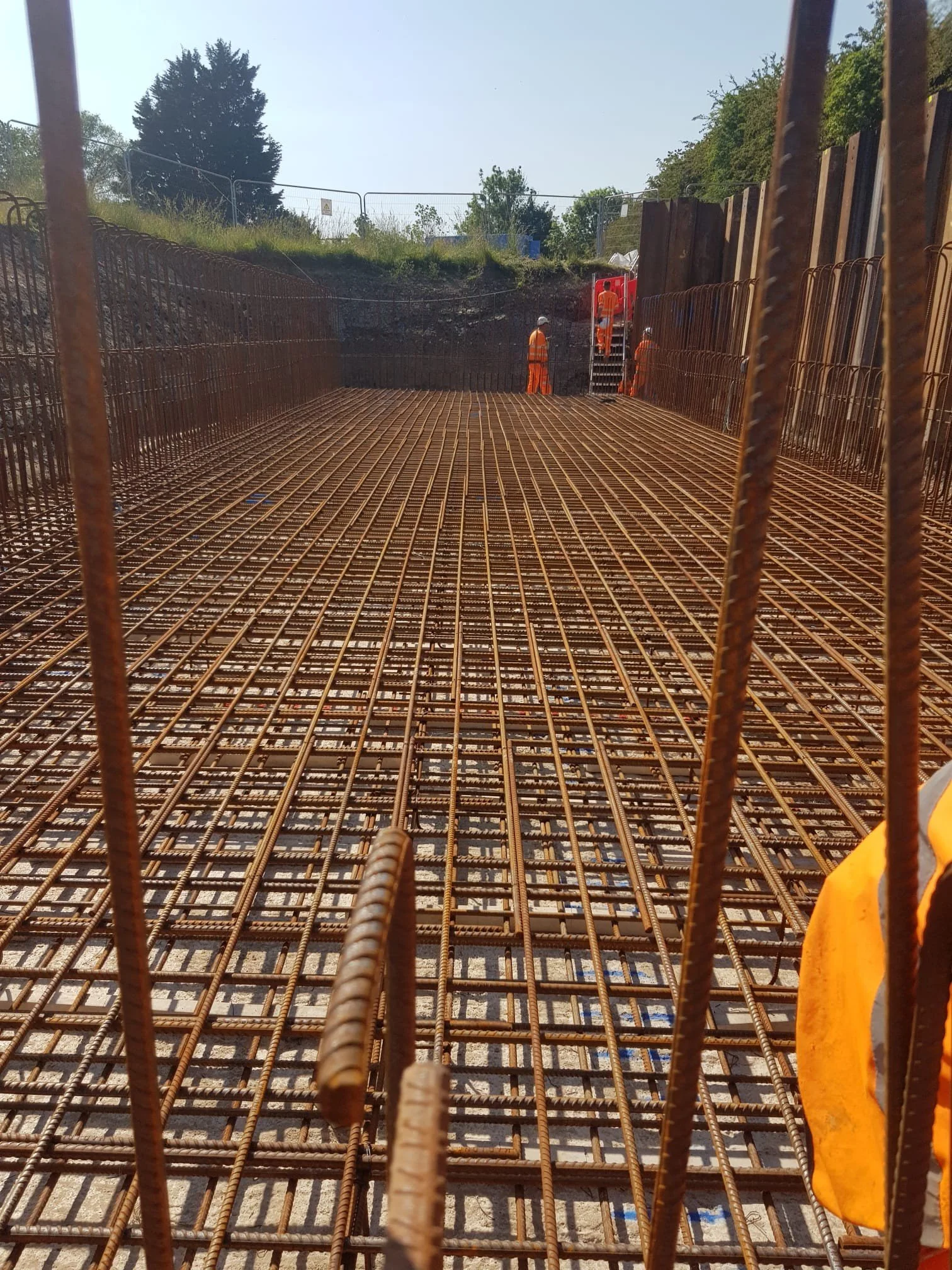 Construction site with steel rebar framework and workers in orange safety gear working near a wooden wall.