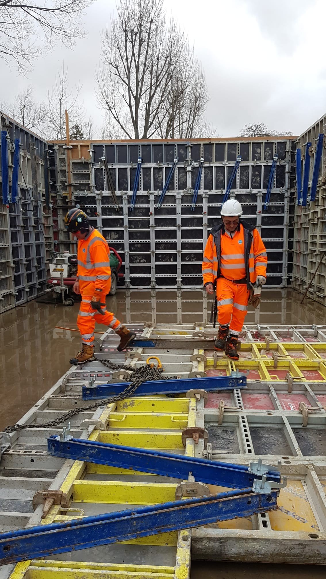 Construction workers in orange safety gear and white helmets working on a construction site with concrete and metal framework.