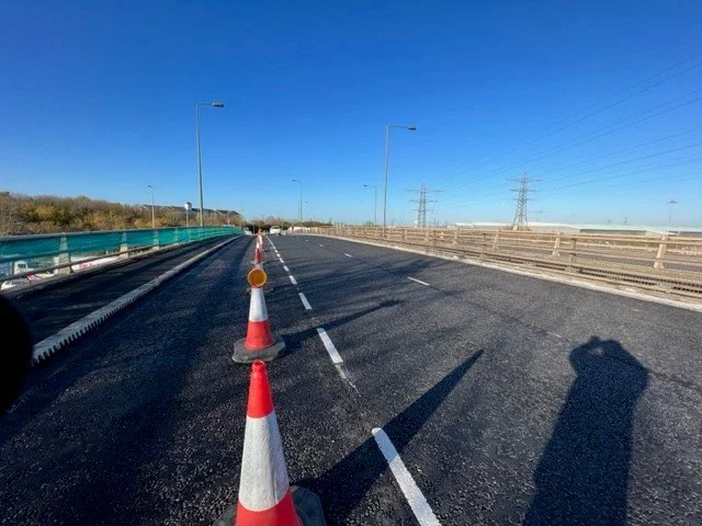 Empty highway lane closed off with orange and white traffic cones, under a clear blue sky with shadows and power lines in the background.