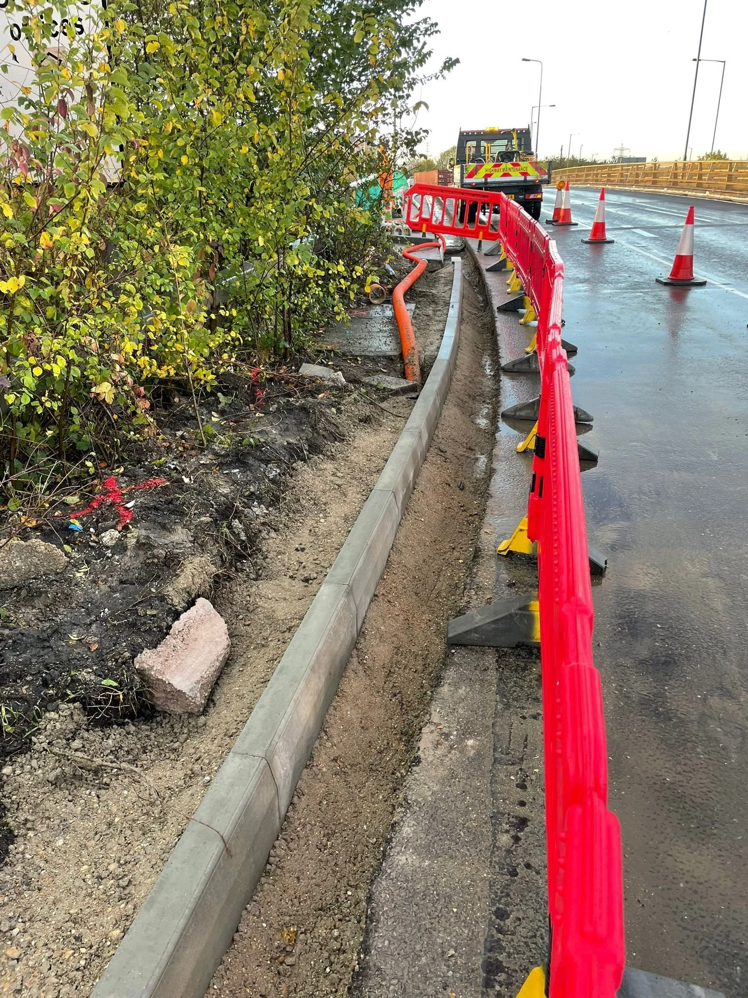 Road construction site with orange barrier fence, orange pipe, construction vehicle, latex band, traffic cones, and a partially paved road next to a grassy area with bushes.