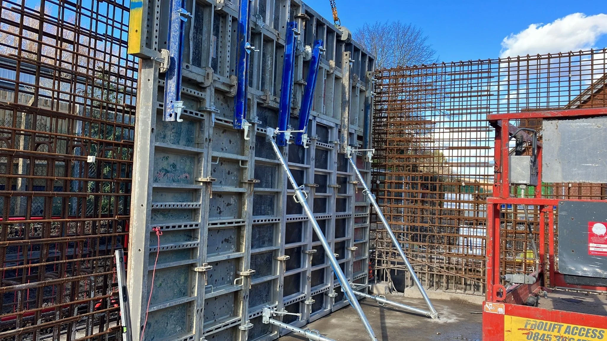 Construction site with reinforcement steel bars and concrete formwork, scaffolding, and safety equipment under a blue sky.