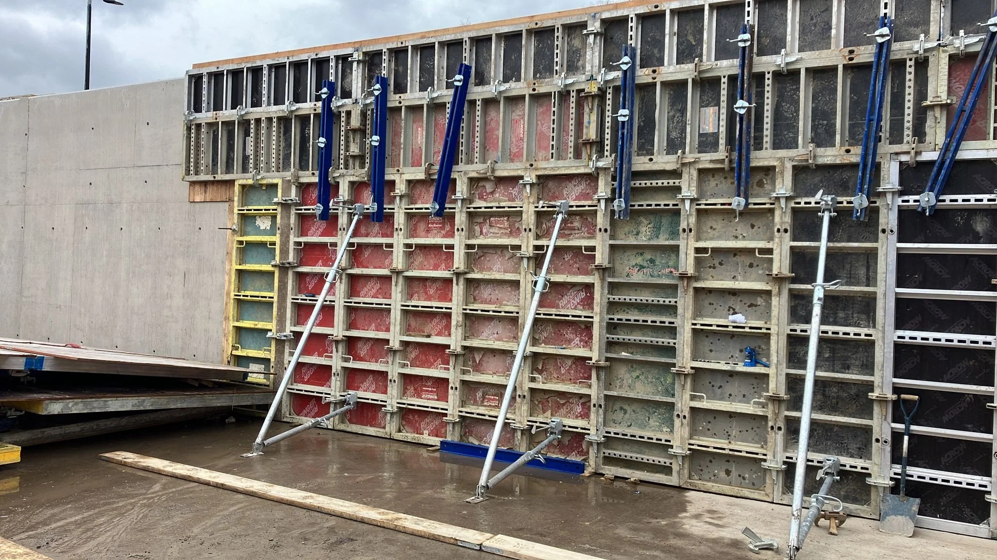 Construction site with temporary wall formwork and scaffolding, wet concrete ground, and construction materials.