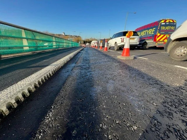 Road construction with orange traffic cones and parked vehicles, including a van and a white car, on a clear day with a blue sky.