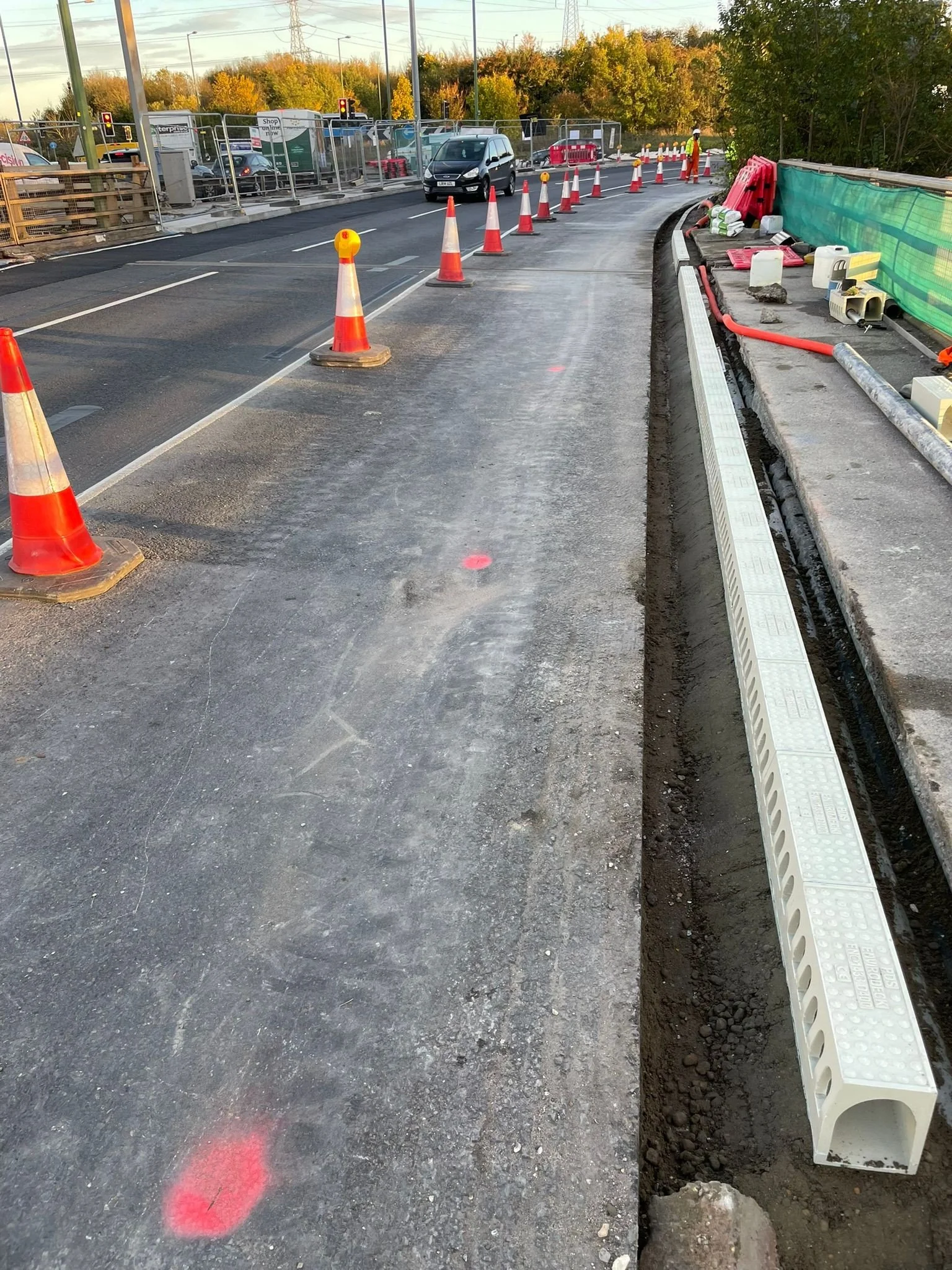 Construction site on a road with orange traffic cones and barriers, with vehicles in the background and construction materials on the side