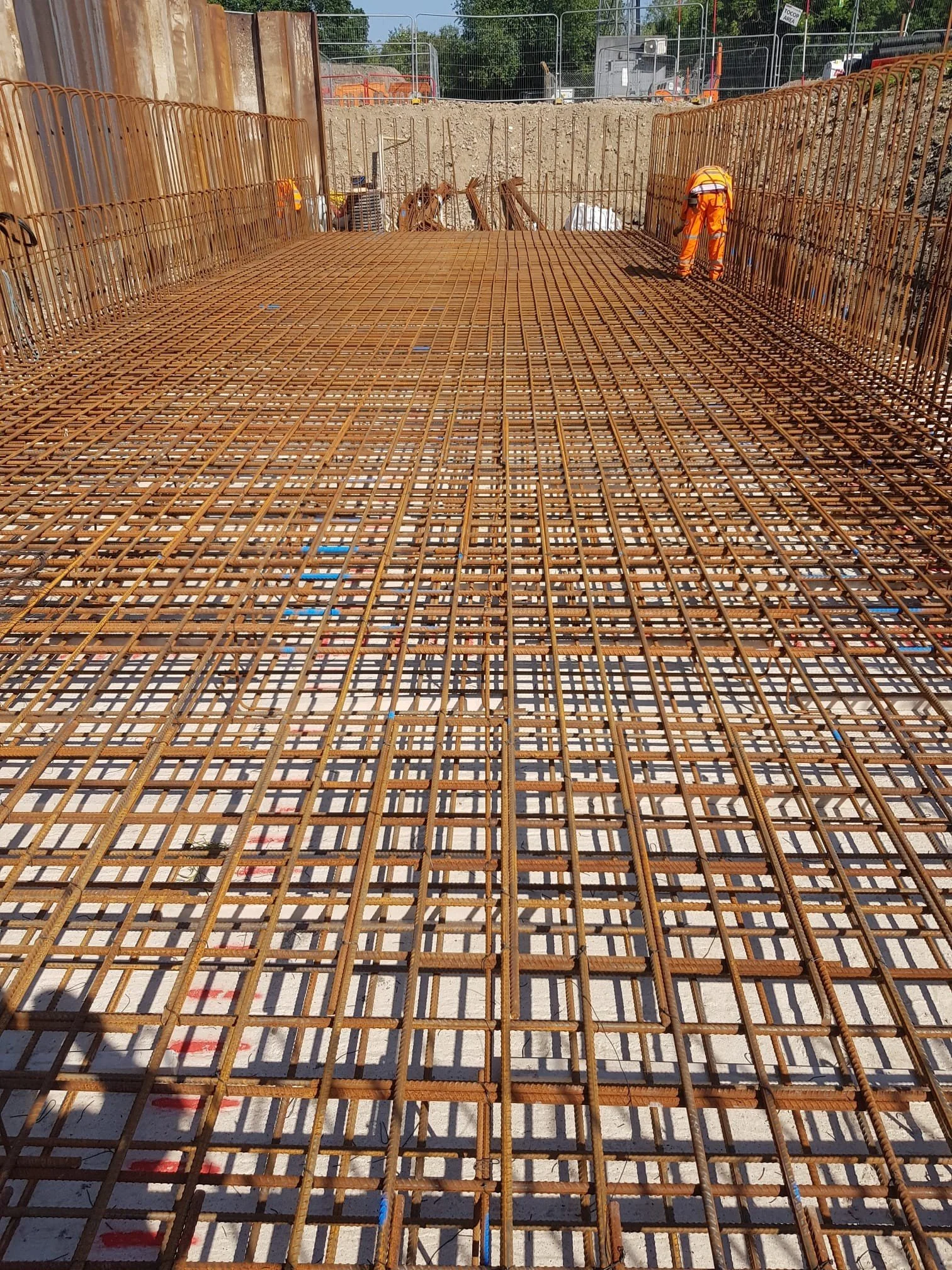 Construction site with steel rebar grid for concrete foundation, worker in orange safety gear working on rebar.