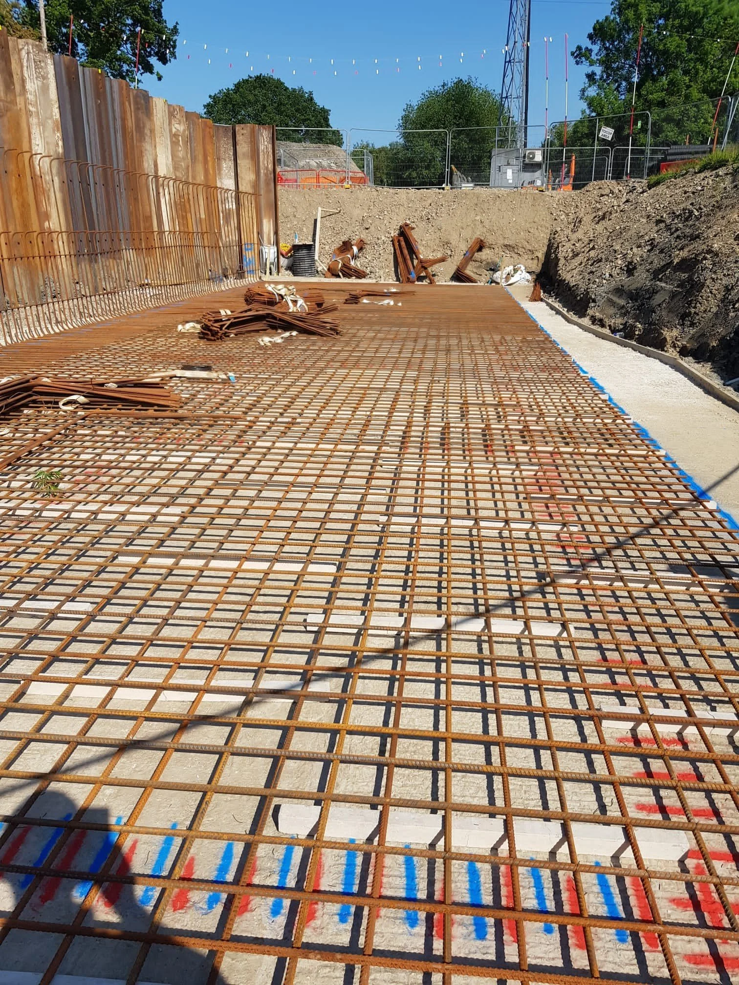 Construction site showing rebar metal grid laid over a concrete foundation for a future structure. Piles of additional rebar are visible in the background, along with construction fencing and equipment, under a clear blue sky.