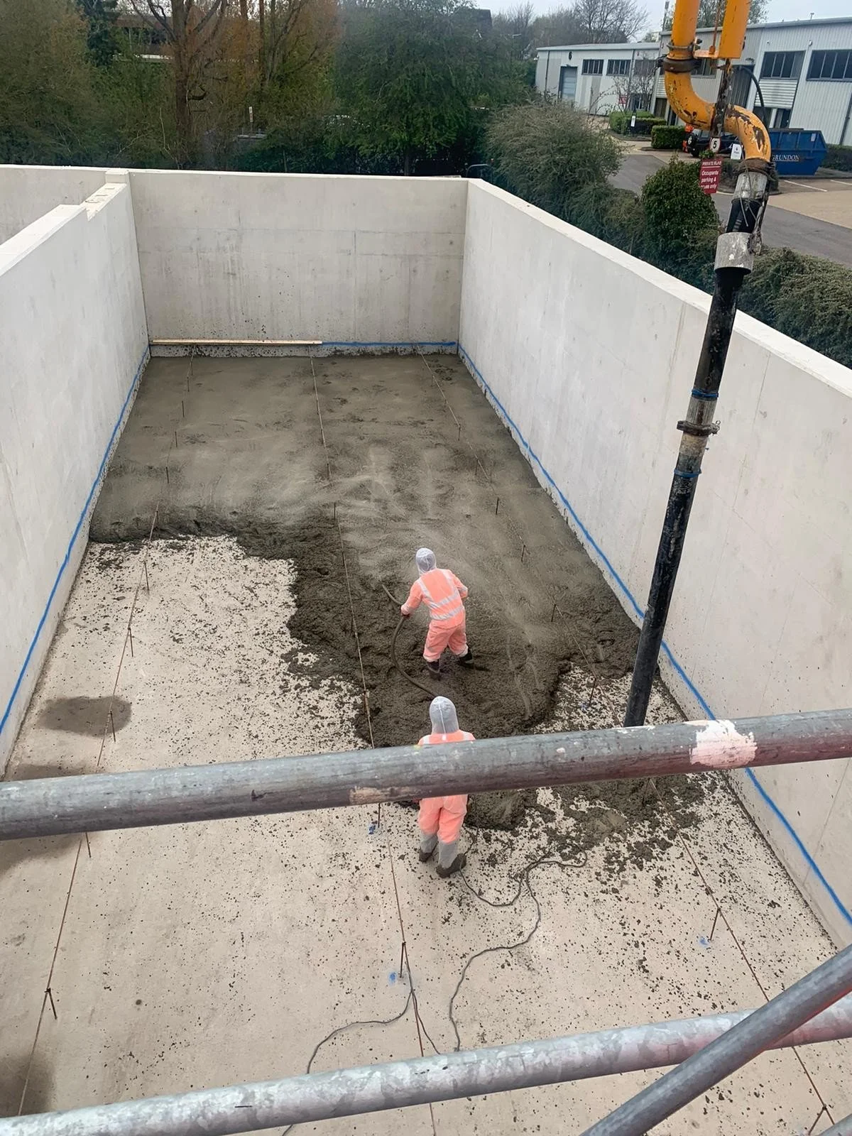 Two construction workers wearing safety suits and helmets work inside a walled area, pouring and spreading concrete on the ground.
