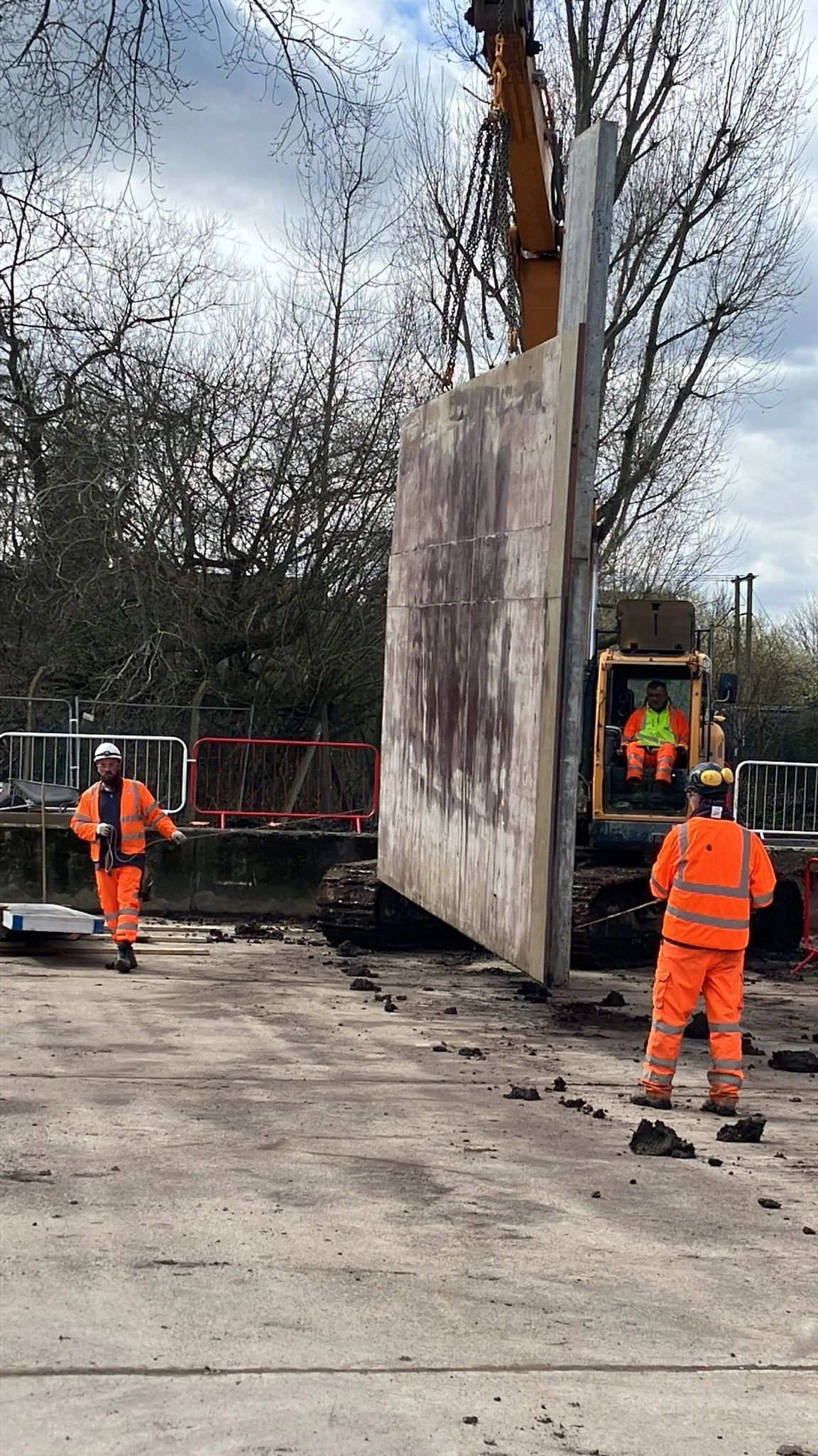 Construction workers wearing orange safety vests and helmets working with a large heavy-duty excavator lifting a concrete wall panel at a construction site.