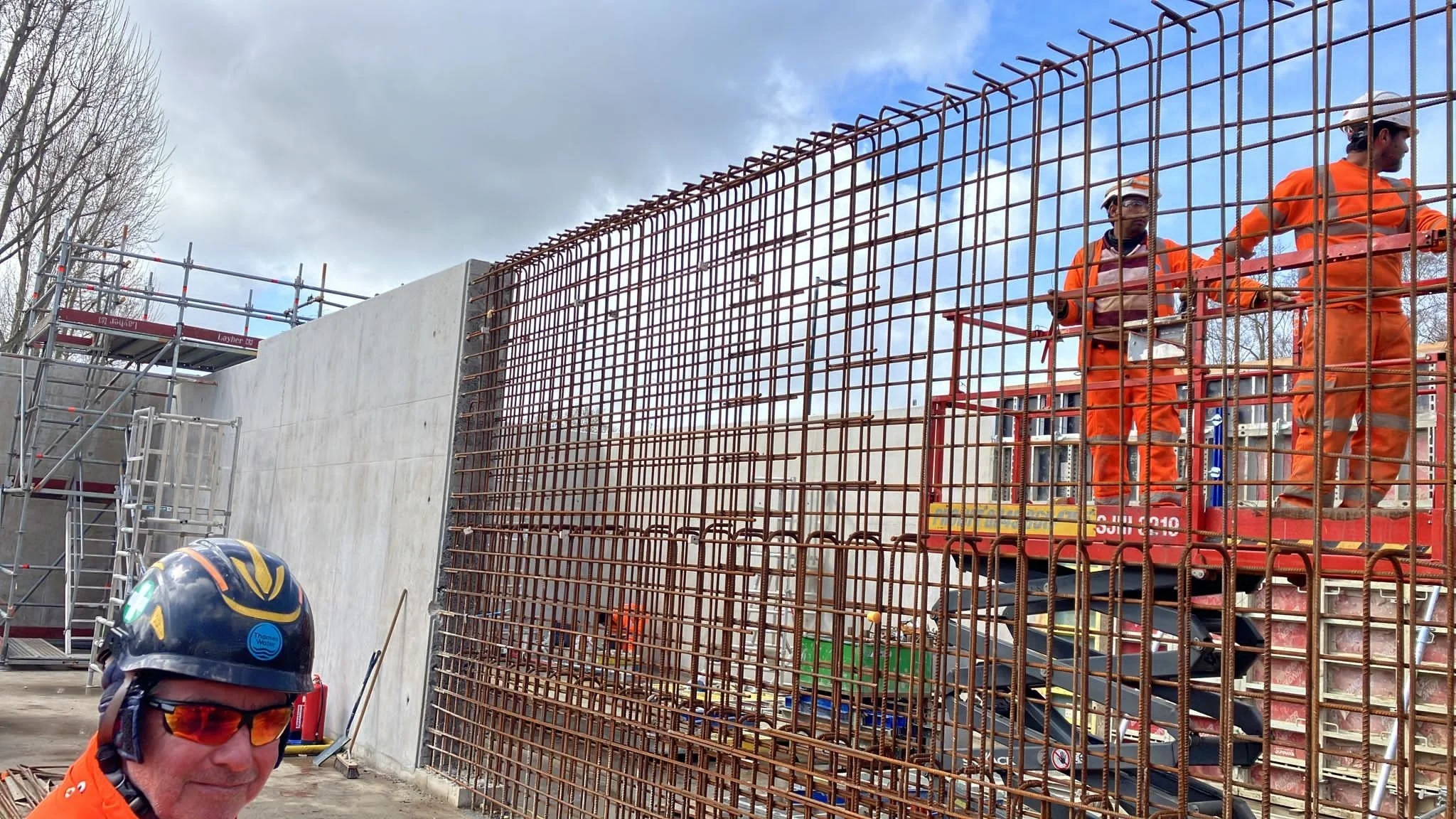 Construction workers in orange safety gear working on a building site, with steel rebar structure and scaffolding visible, and a man in the foreground wearing a helmet and sunglasses.