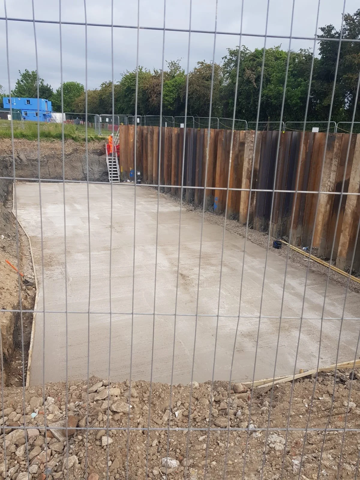View of a construction site with freshly poured concrete slab, surrounded by a temporary metal fence, with soil and rocks at the edges and a rusted metal wall at the back, with trees and cloudy sky in the background.