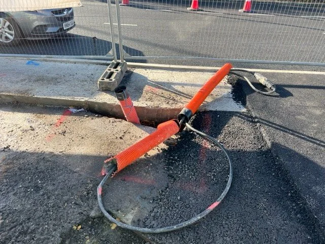 A construction site with orange pipes and a hose on a pavement, surrounded by a temporary metal fence.