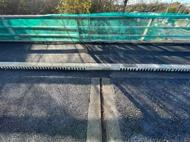 Close-up of a railway track embedded in asphalt on a road, with a green construction barrier and trees in the background.