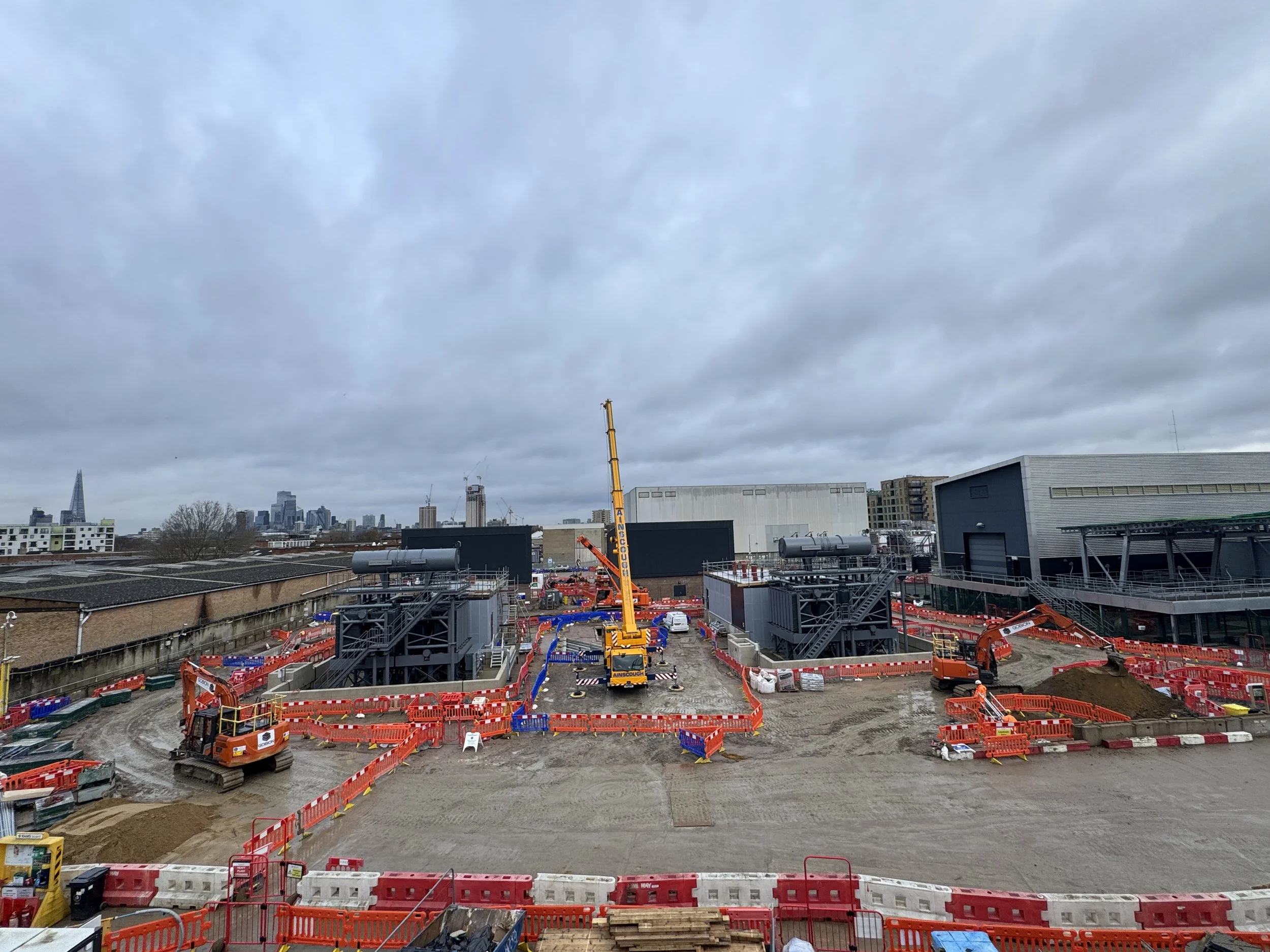 Construction site with heavy machinery, orange safety barriers, and cloudy sky in an urban area.