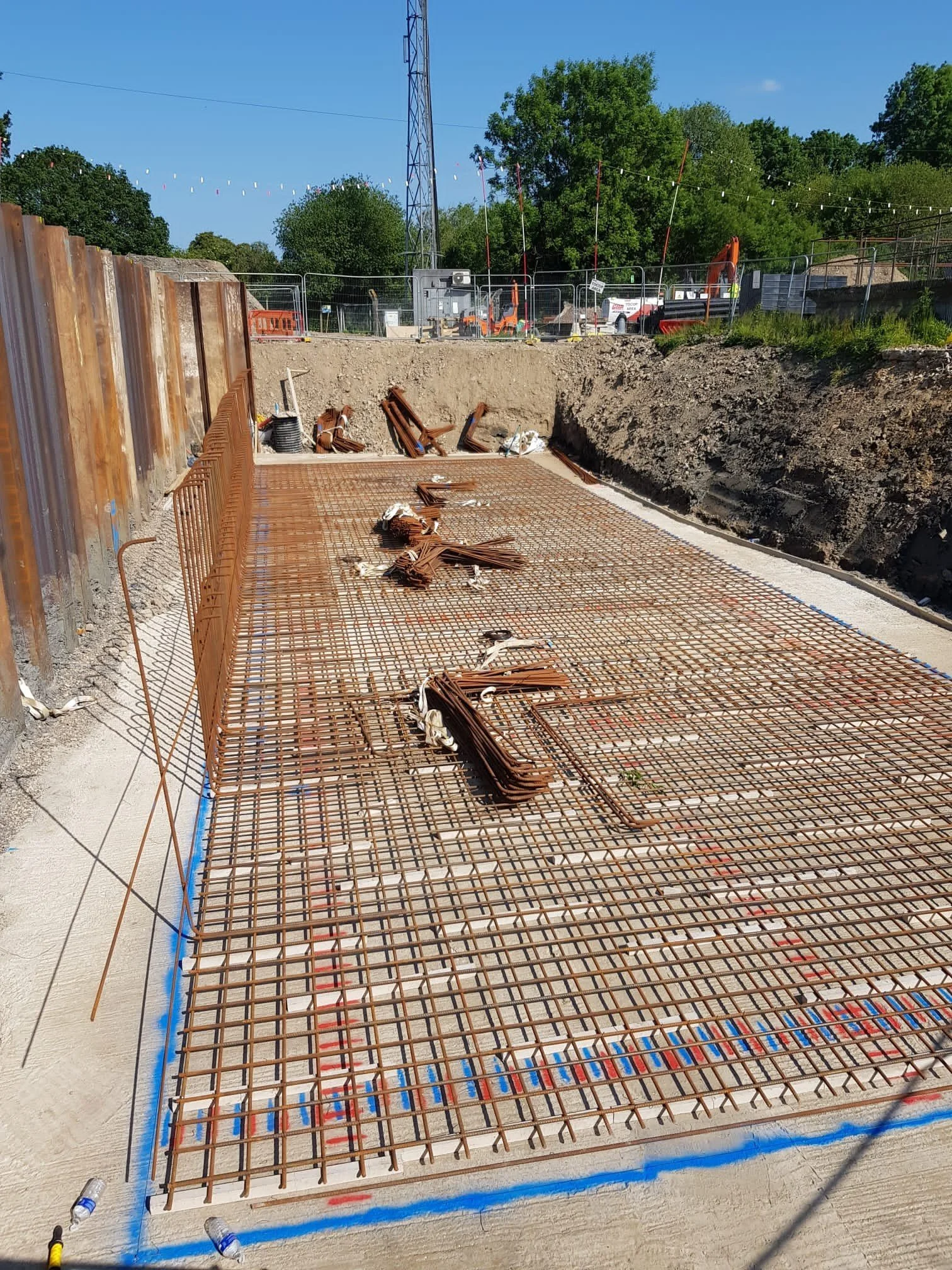 Construction site with rebar grid preparing for concrete pour, surrounded by dirt walls and construction equipment in the background under a blue sky.