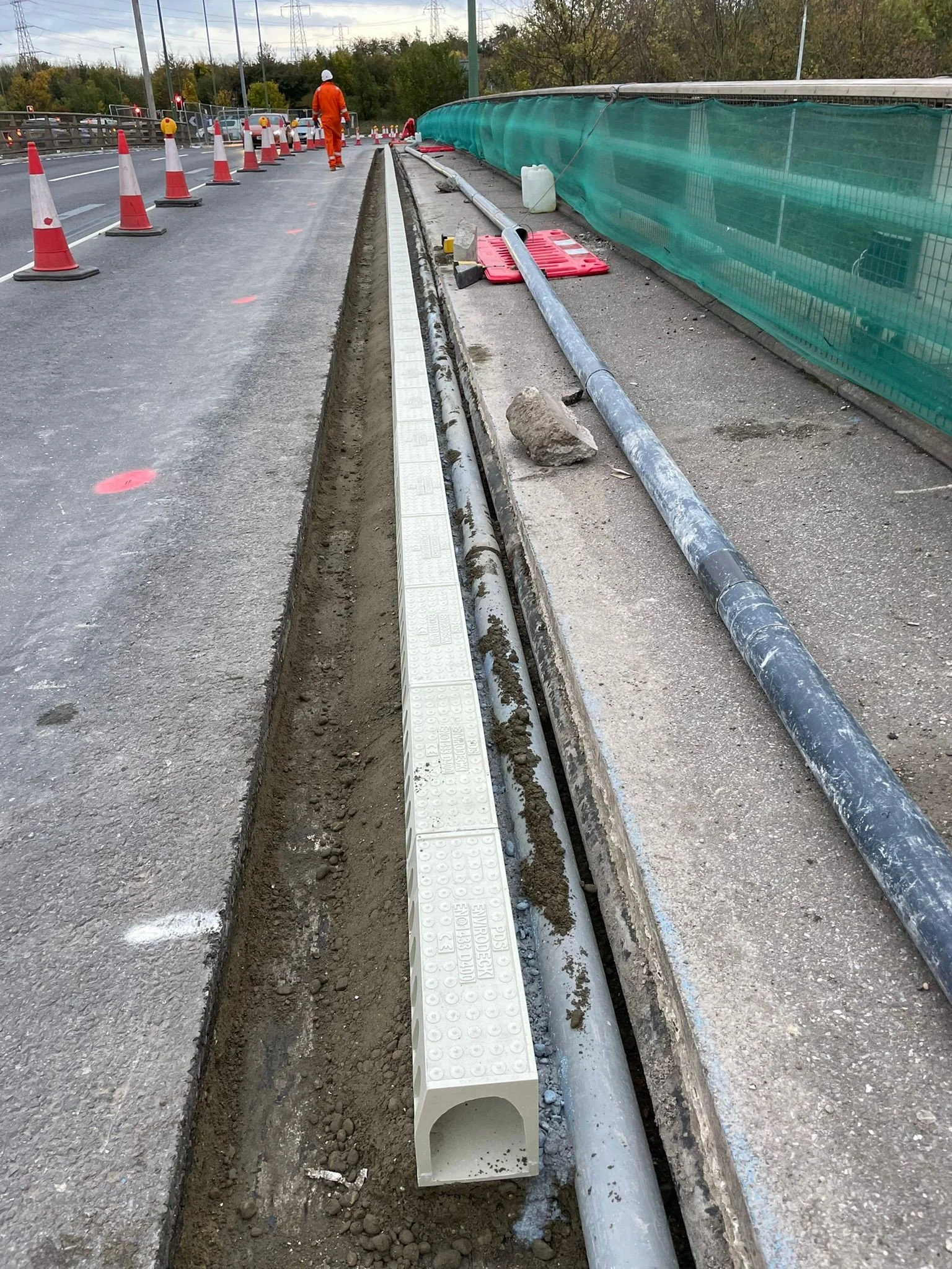 Construction worker working on sidewalk irrigation system with pipes, traffic cones, and safety barriers on the road.