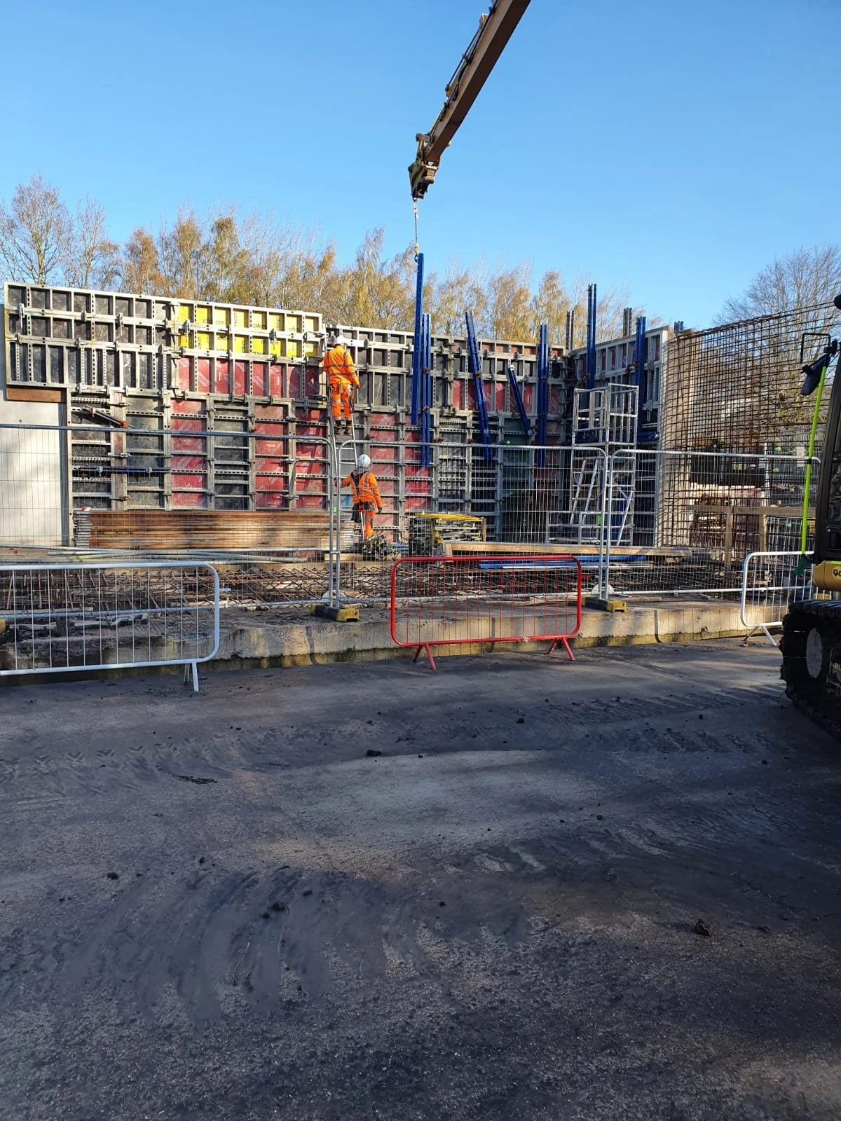 Construction workers in orange safety uniforms working on a construction site with a large concrete wall and scaffolding, surrounded by safety barriers.