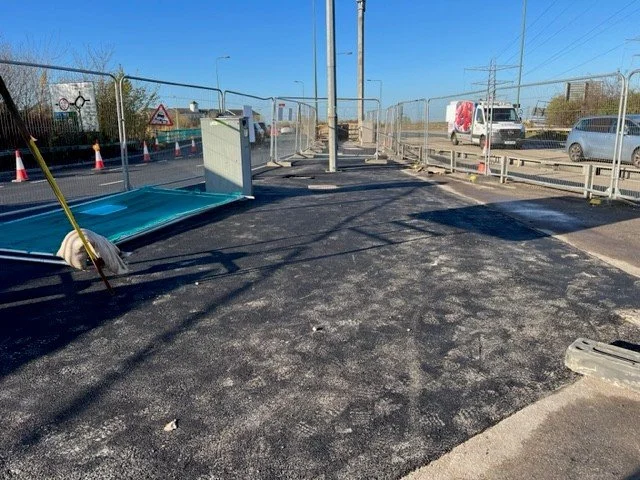 A construction site on a road with asphalt pavement, traffic cones, and fencing. There is construction equipment like a broom and a small structure, with cars and power lines in the background under a clear blue sky.