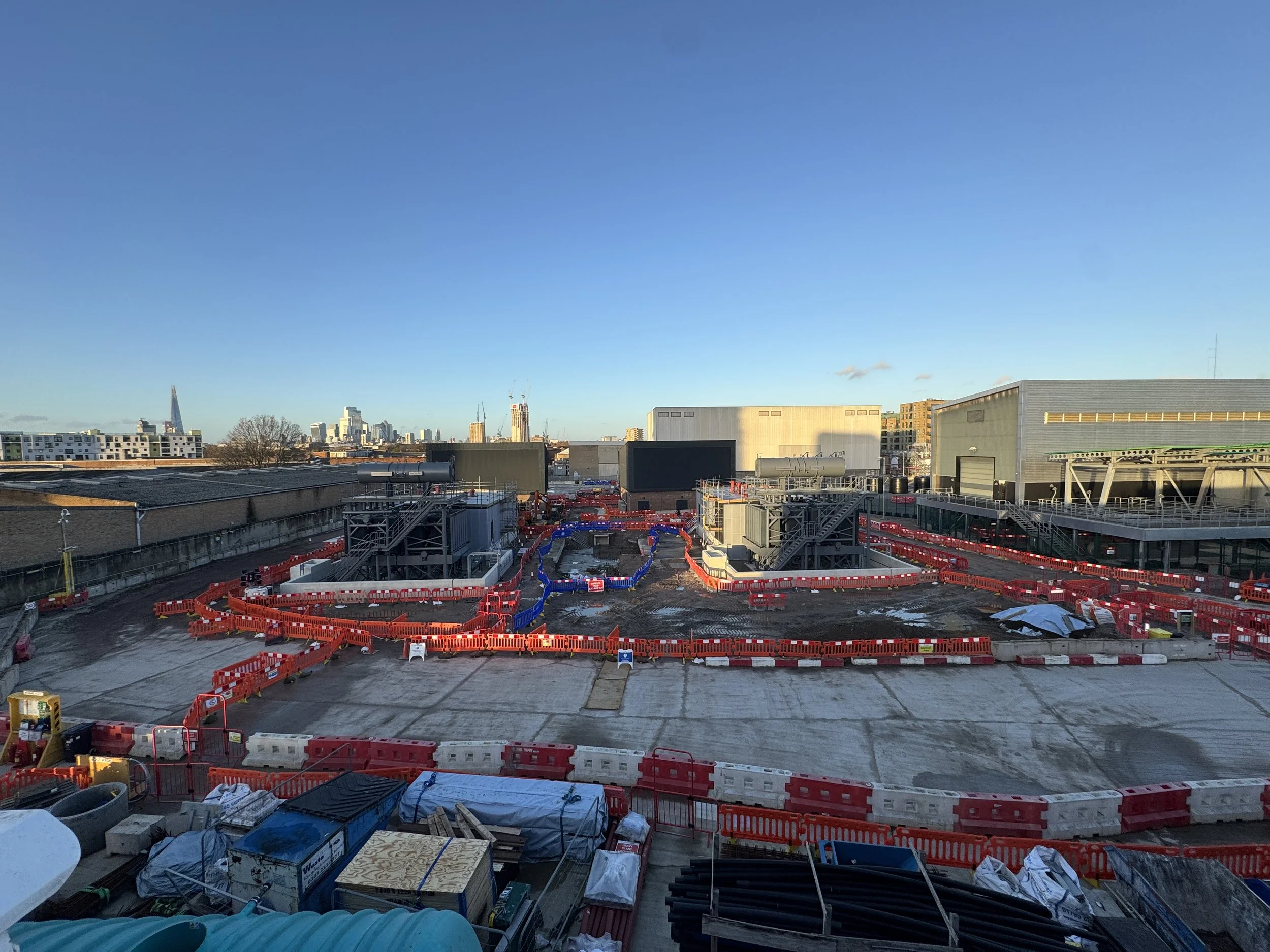 Construction site with orange safety barriers, equipment, and building structures in a cityscape under a clear blue sky.