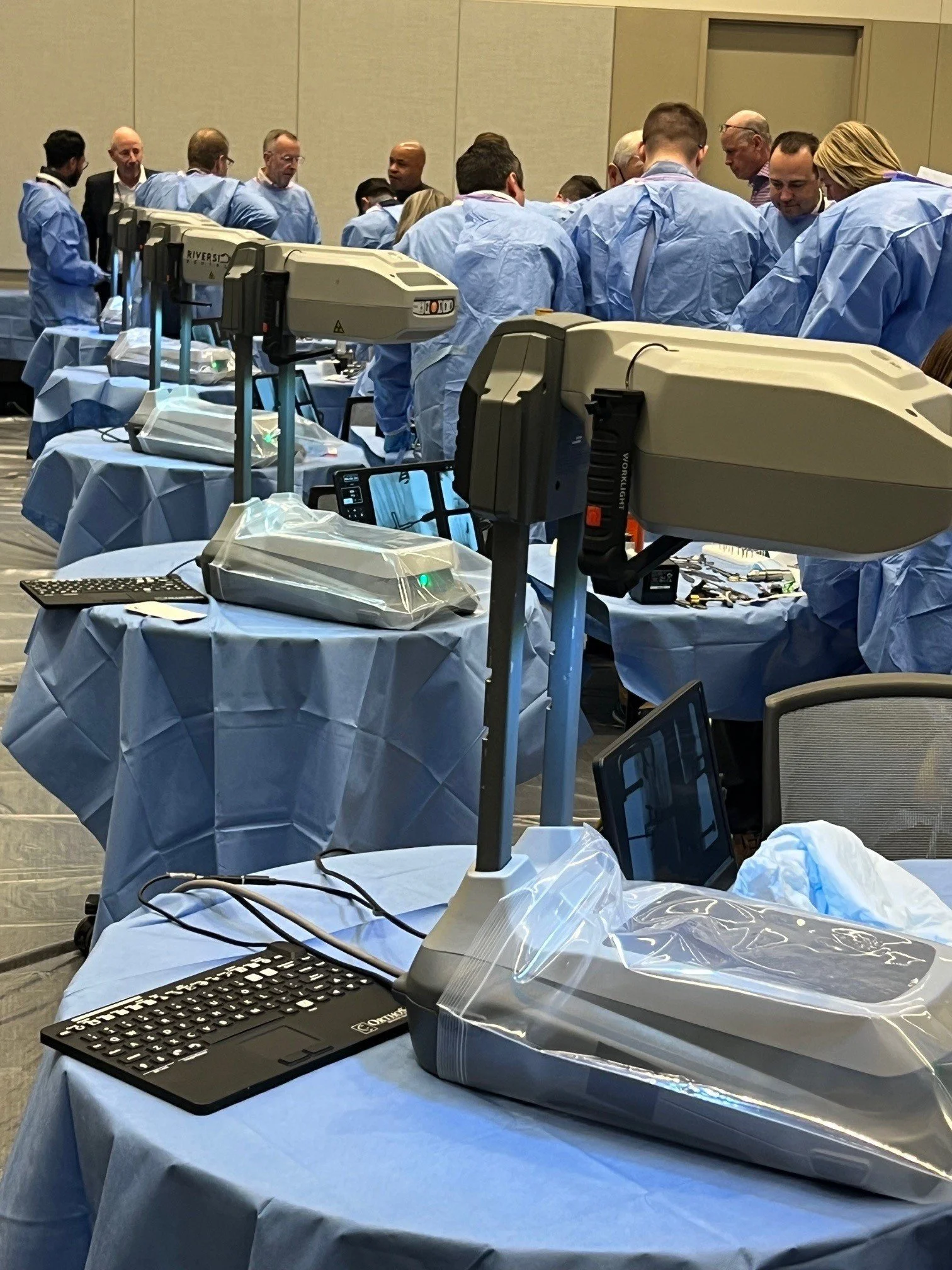 Medical professionals wearing blue scrubs gathered behind tables with medical equipment, monitors, and supplies in a conference or training setting.