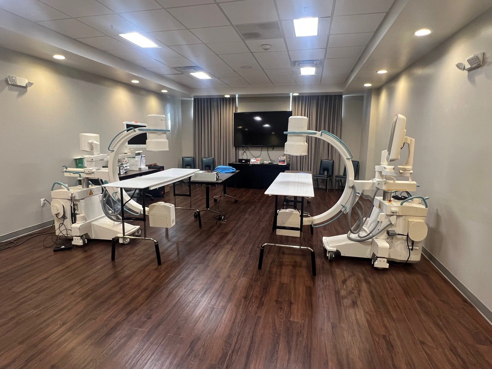 Medical room set up for imaging procedures with two large, C-arm X-ray machines, tables, chairs, and electronic equipment.