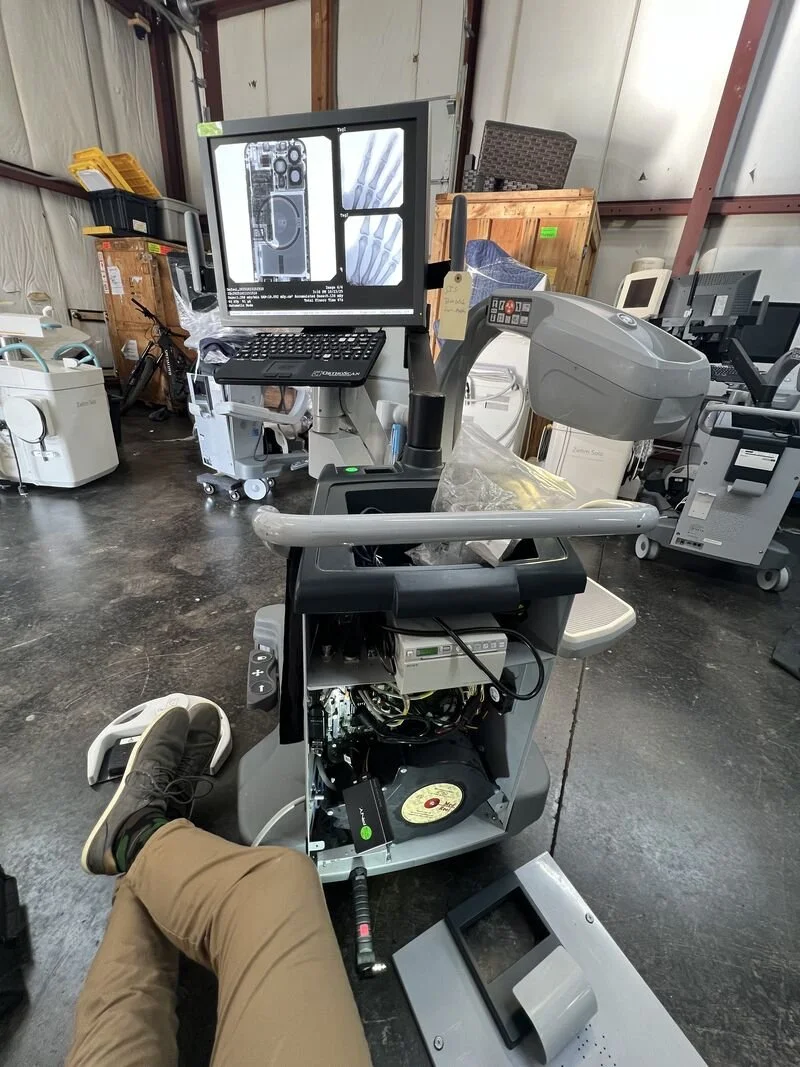 Medical imaging equipment in a warehouse, including an X-ray machine showing hand and chest images on the monitor, with a person's leg and shoe visible in the foreground.