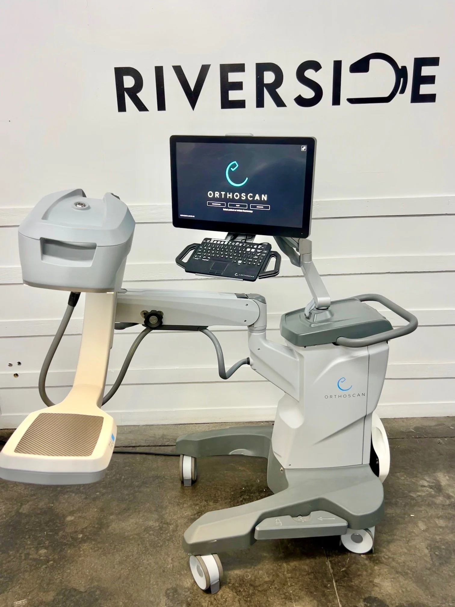 Medical imaging machine with a monitor displaying 'ORTHOSCAN' in a clinical setting with the word 'RIVERSIDE' on the wall.