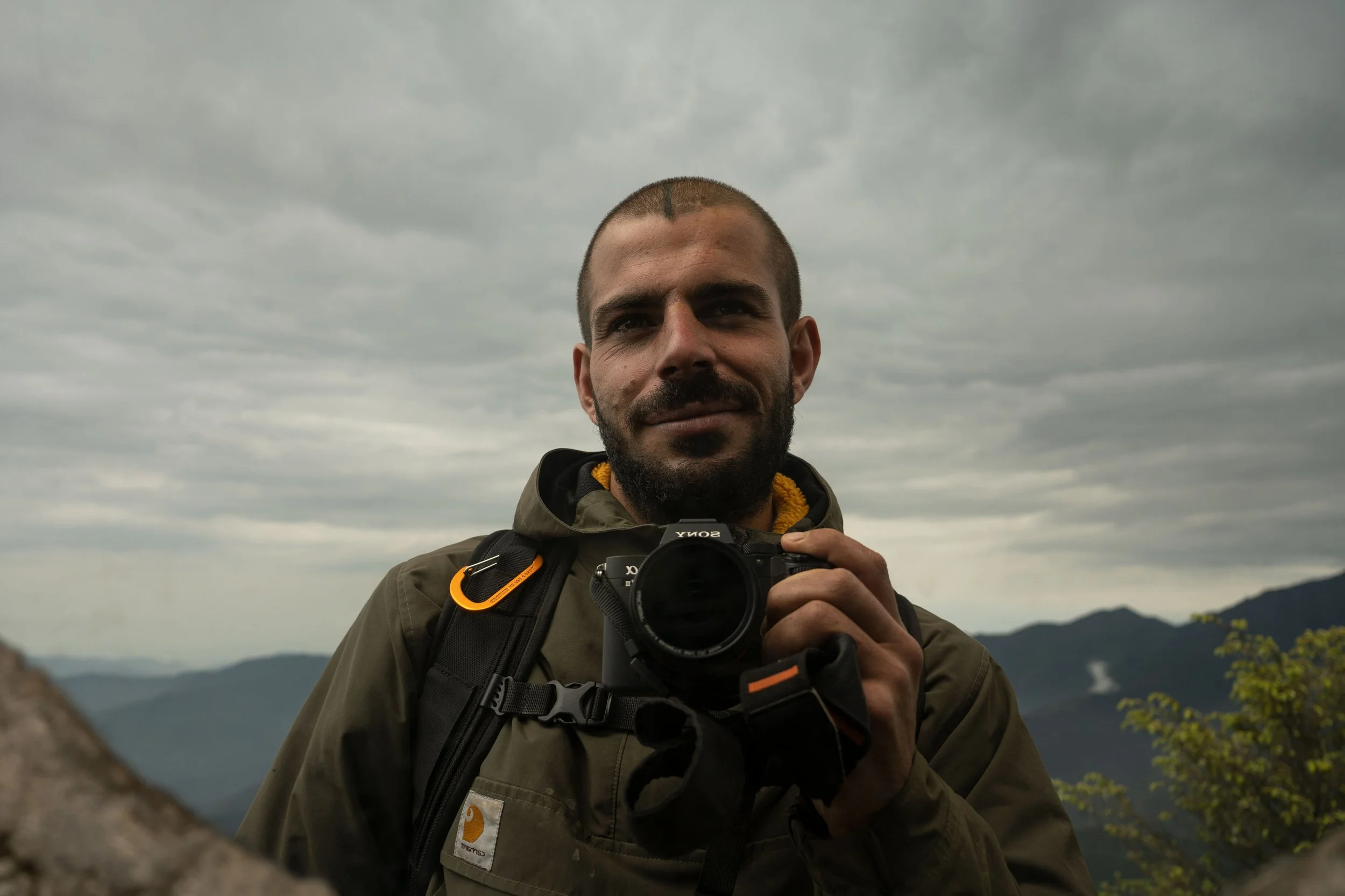 Un hombre con barba y cabello corto, usando chaqueta de color oliva y mochila, sosteniendo una cámara réflex Sony, en un paisaje montañoso con cielo nublado.