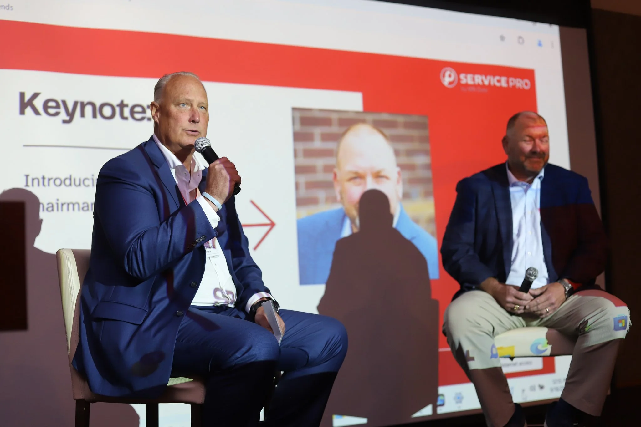 Two men sitting on chairs, participating in a panel discussion at a conference. Behind them is a large screen displaying a presentation slide with the title 'Keynote' and a red logo for ServicePro. The man on the left, dressed in a blue suit, is speaking into a handheld microphone. The man on the right, dressed in a dark blazer and beige pants, is holding a microphone in his lap.