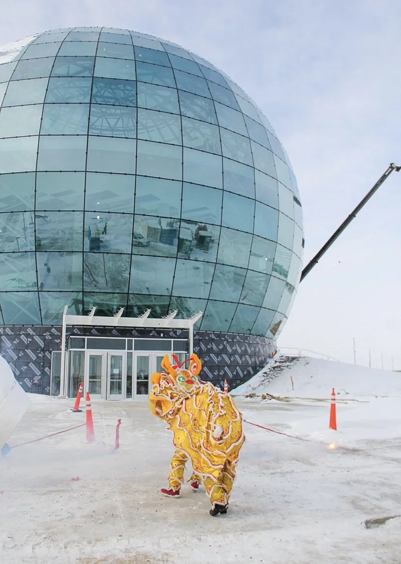 A person wearing a colorful lion dance costume standing in front of a large glass spherical building on a snowy day.