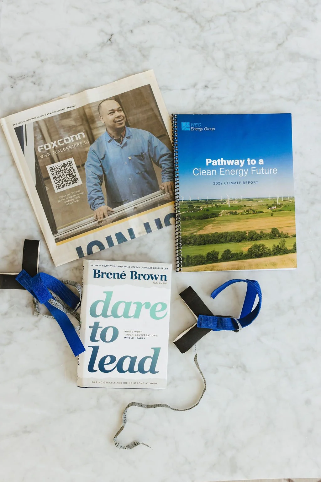 A magazine, a spiral-bound report, a book, and two lanyards with straps are arranged on a white marble surface. The magazine features a man smiling and wearing a blue work shirt, and has a QR code on the cover. The report is titled 'Pathway to a Clean Energy Future,' and the book is titled 'Dare to Lead' by Brené Brown.
