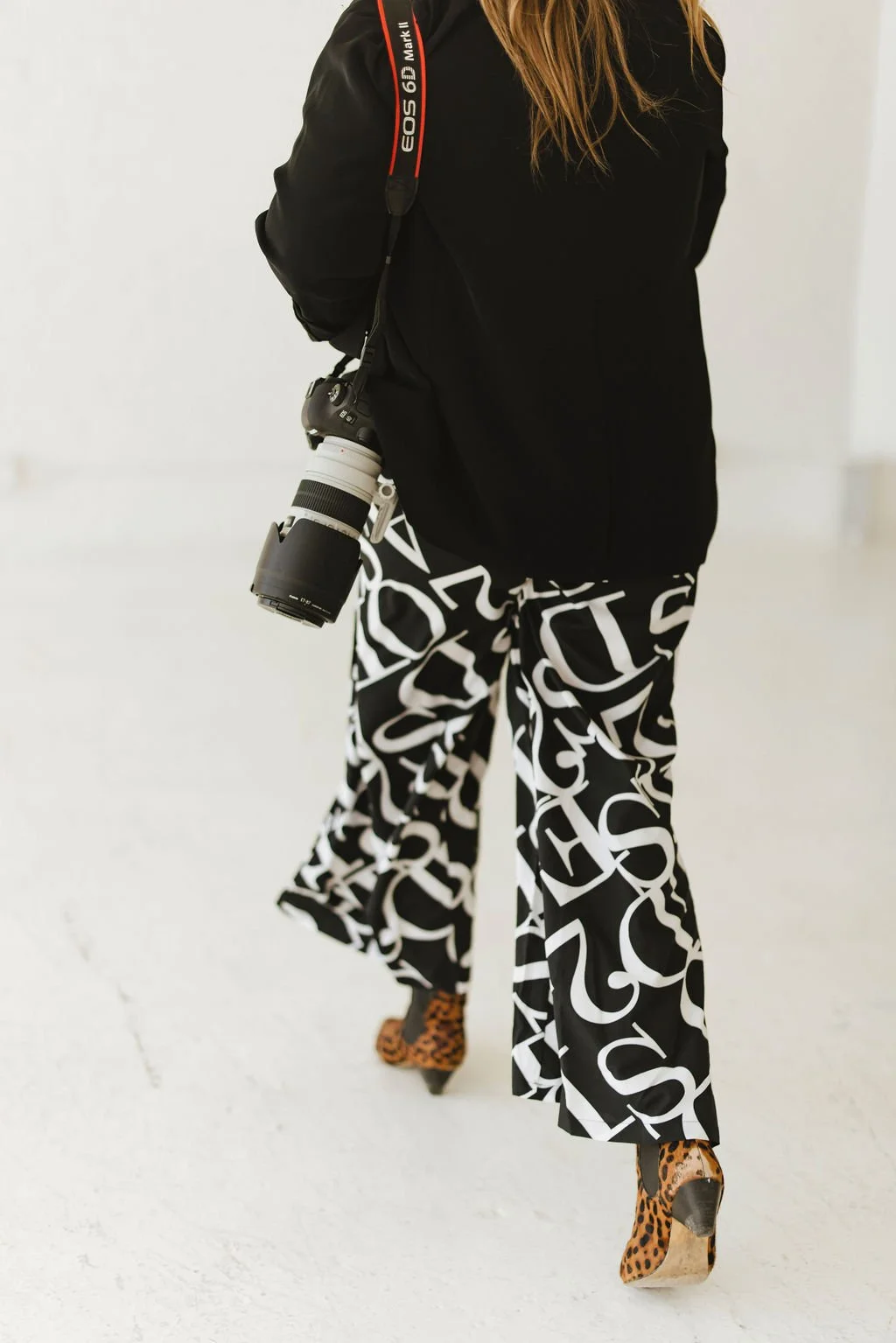 Alyce Peterson with a professional camera hanging from her neck, wearing patterned wide-leg pants with large white lettering and leopard-print high heels, standing in a minimalist white room.