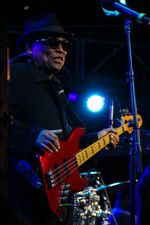 A man wearing a black hat, sunglasses, and a black jacket playing a red electric bass guitar on stage with blue and white stage lighting.