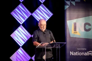An older man with glasses standing at a clear podium, speaking on a stage with a black background and decorative purple geometric shapes, in front of a large screen that has the abbreviation 'LNC' and a logo for National Memorial.