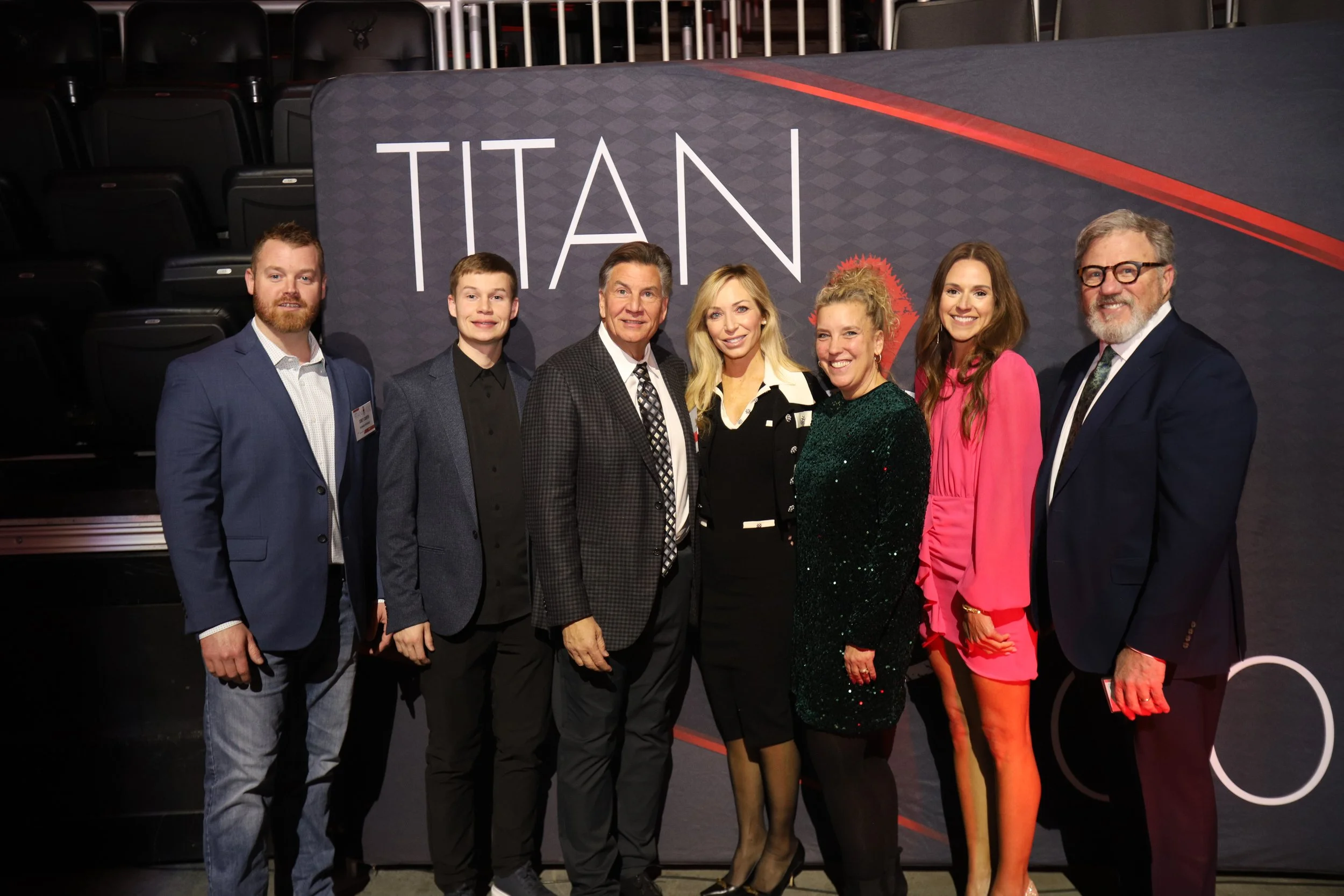 A group of eight people posing for a Titan 100 photo in front of a backdrop with the word 'TITAN' on it. They are dressed in business or semi-formal attire and are standing indoors, likely at an event or premiere.