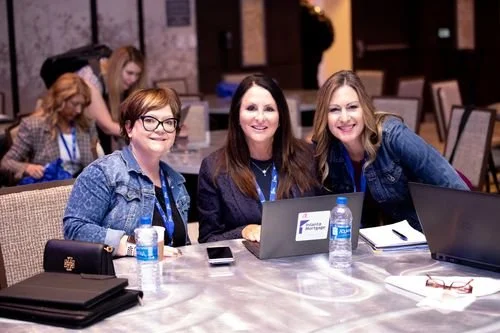 Three women sitting at a conference table with laptops and water bottles, smiling at the camera at a professional event.