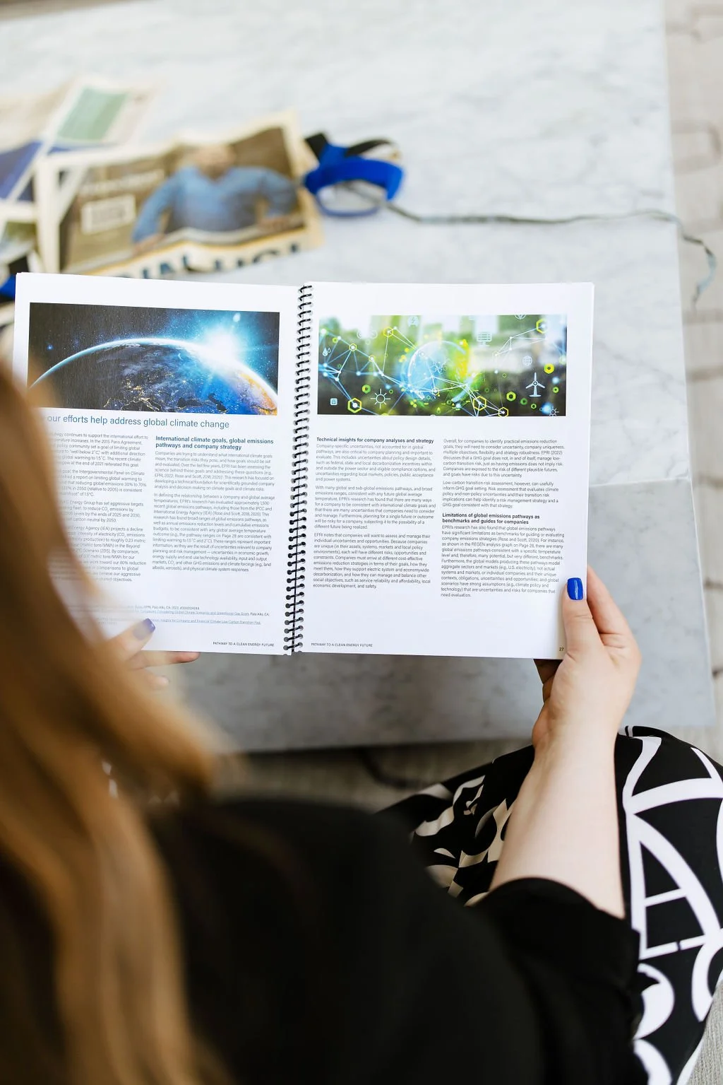 A person holding an open booklet with two pages of infographics and text about climate change and technology. On the table in the background are some magazines and a pair of blue headphones.