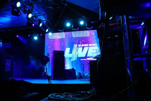 Stage setup with a large screen displaying the Super Bowl LIV logo, surrounded by stage lights, equipment, and cables.