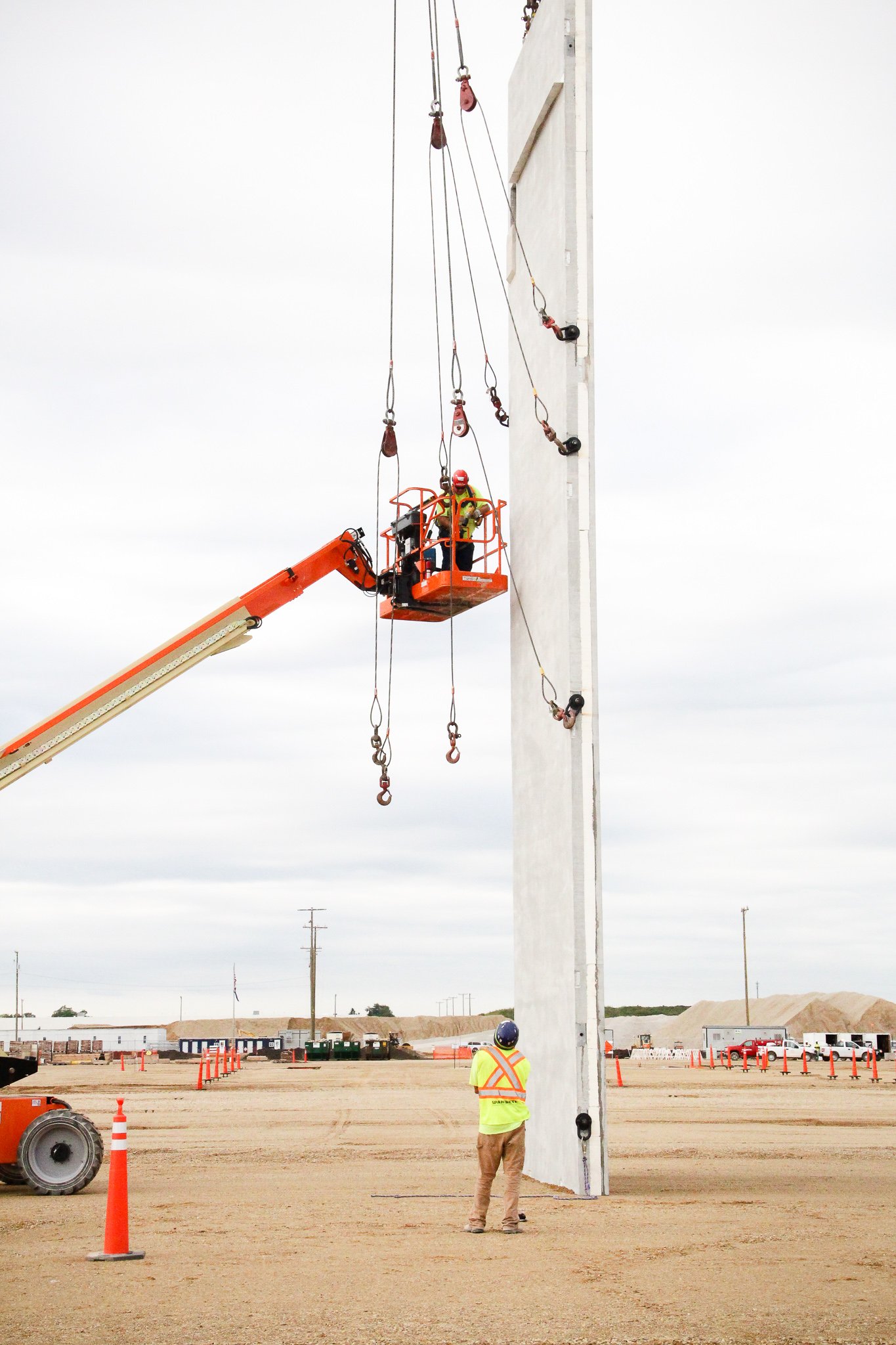 Construction workers using a lift to install or inspect large vertical structure at a construction site with orange safety cones and machinery in the background.