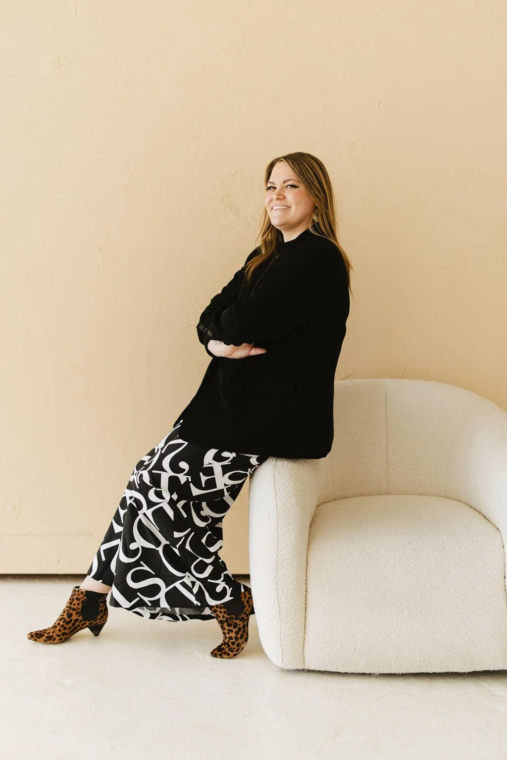 A woman sitting on the edge of a light-colored armchair, wearing a black blazer, black and white patterned pants, and leopard print boots, smiling with arms crossed, against a beige wall.