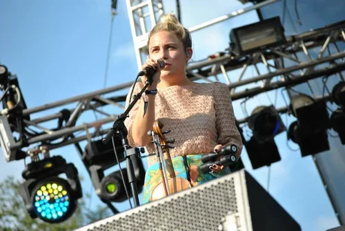 A woman at Lollapalooza performing on stage with a microphone, outdoors, under a clear blue sky, with stage lights and scaffolding.