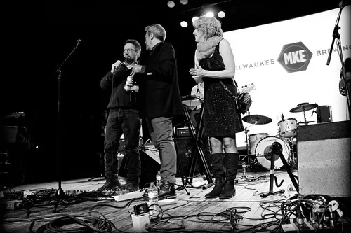 Three people on stage, two men and one woman, engaged in conversation with microphones, set up with musical instruments and equipment, with a backdrop displaying the Milwaukee Bucks logo and event branding.