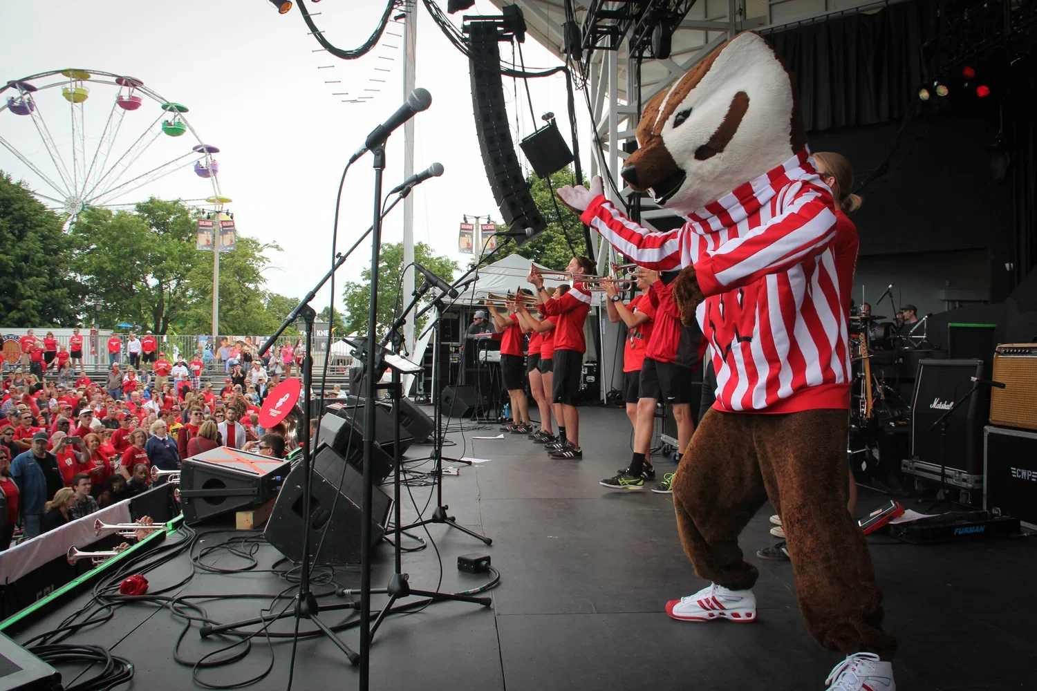 A UW Madison mascot costume with red and white striped shirt performing on stage at an outdoor concert, with a band playing and a crowd of fans, many in red shirts, in front of the stage. A Ferris wheel is visible in the background.