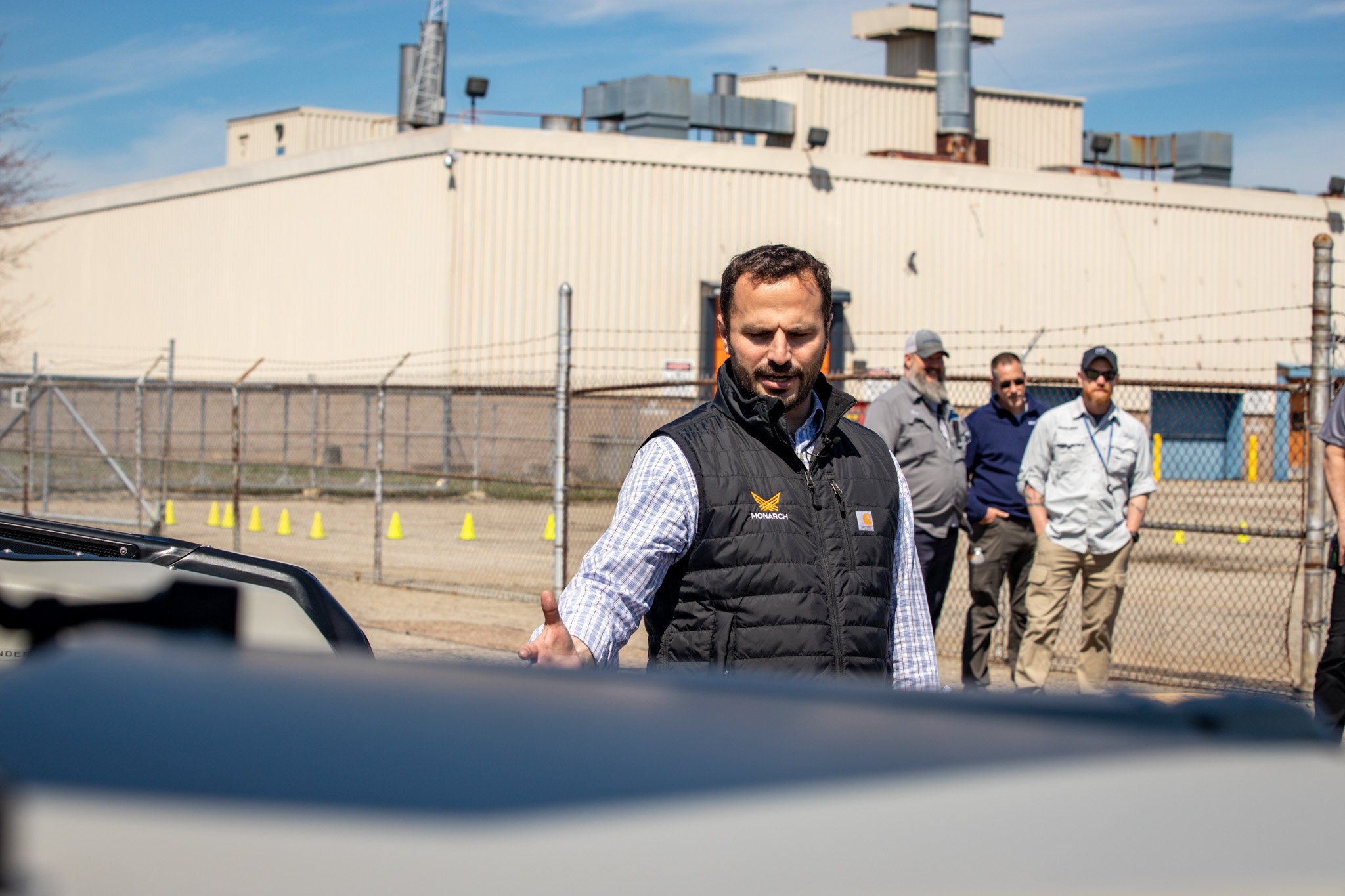 A man in a black vest speaking or gesturing outdoors with a group of men in the background behind a chain-link fence and industrial building, under a blue sky.