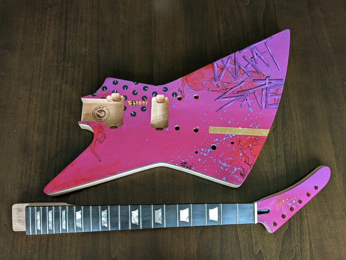 A guitarwith a pink and purple painted design, featuring black spots, blue splatters, and text.