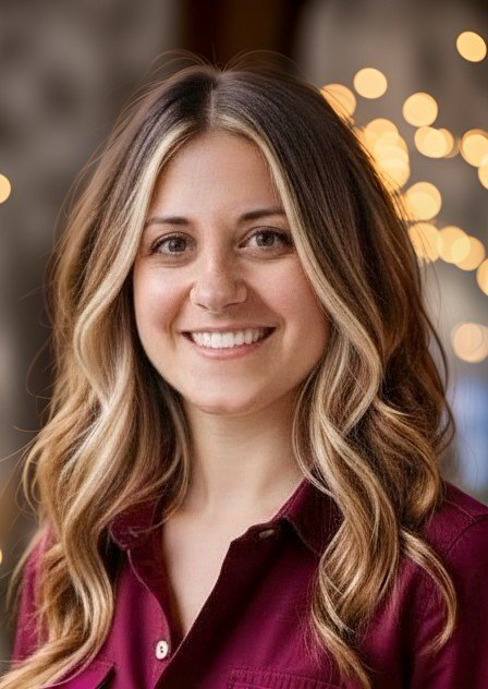 A young woman with long, wavy blonde hair smiling at the camera, wearing a maroon shirt, with blurred lights in the background.