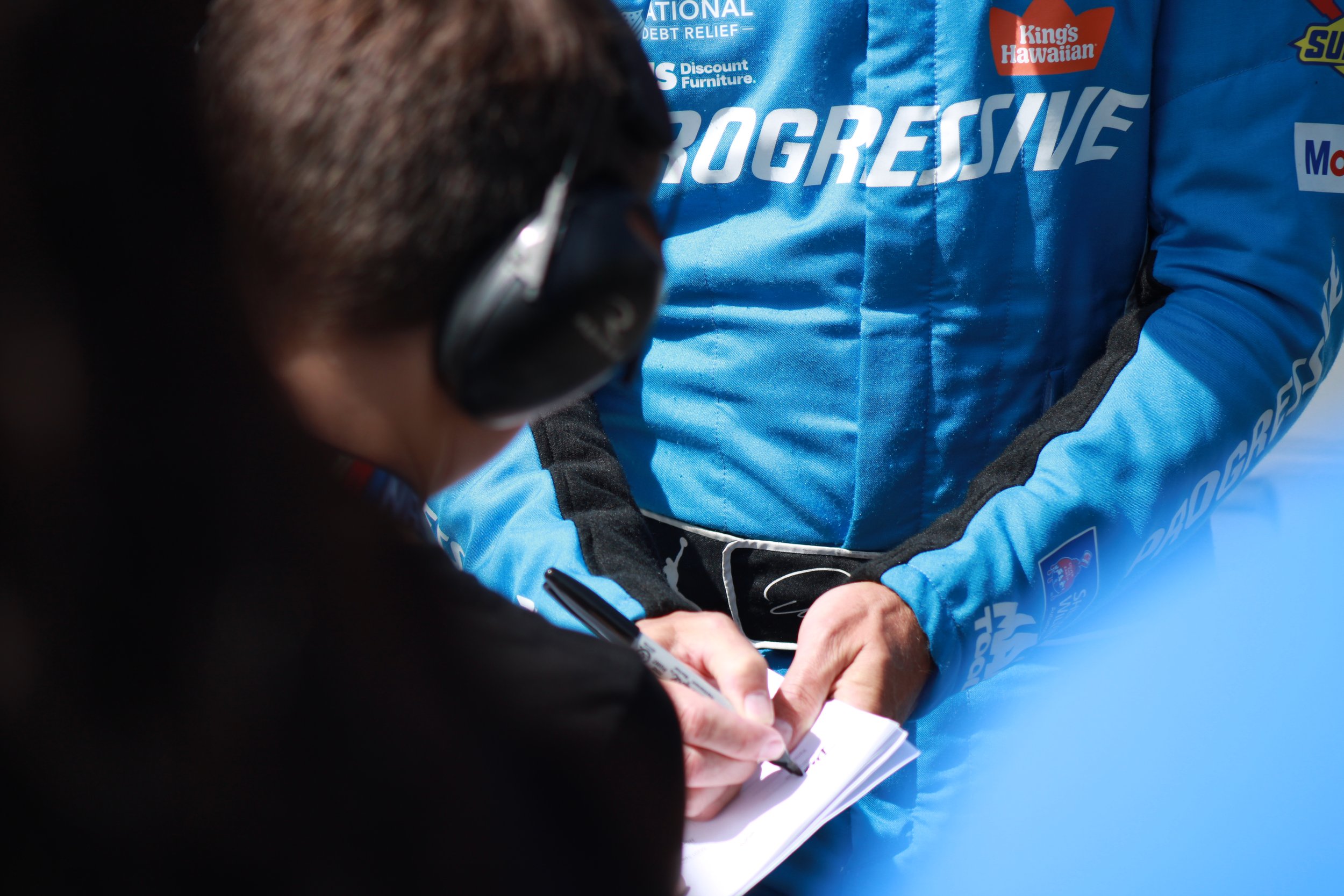 A person wearing black headphones writes on a notepad, next to a man in a blue racing suit with sponsor logos.