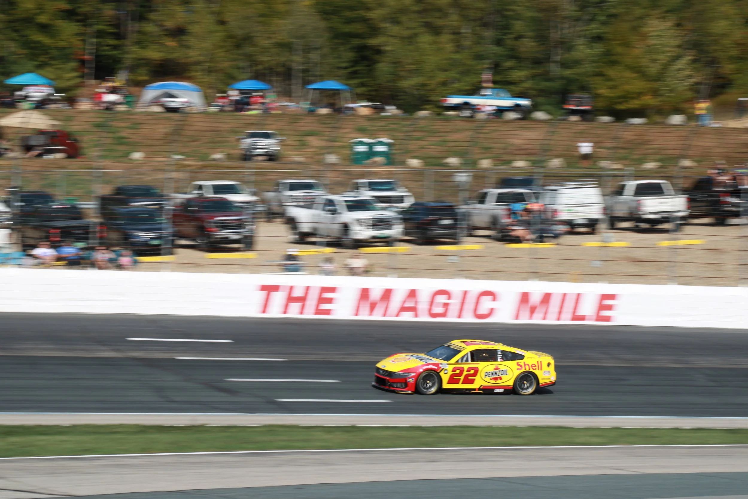 A yellow race car with red and black accents, bearing the number 22, speeding on a racetrack with a blurred background of cars and spectators, with the text 'THE MAGIC MILE' visible on the wall.