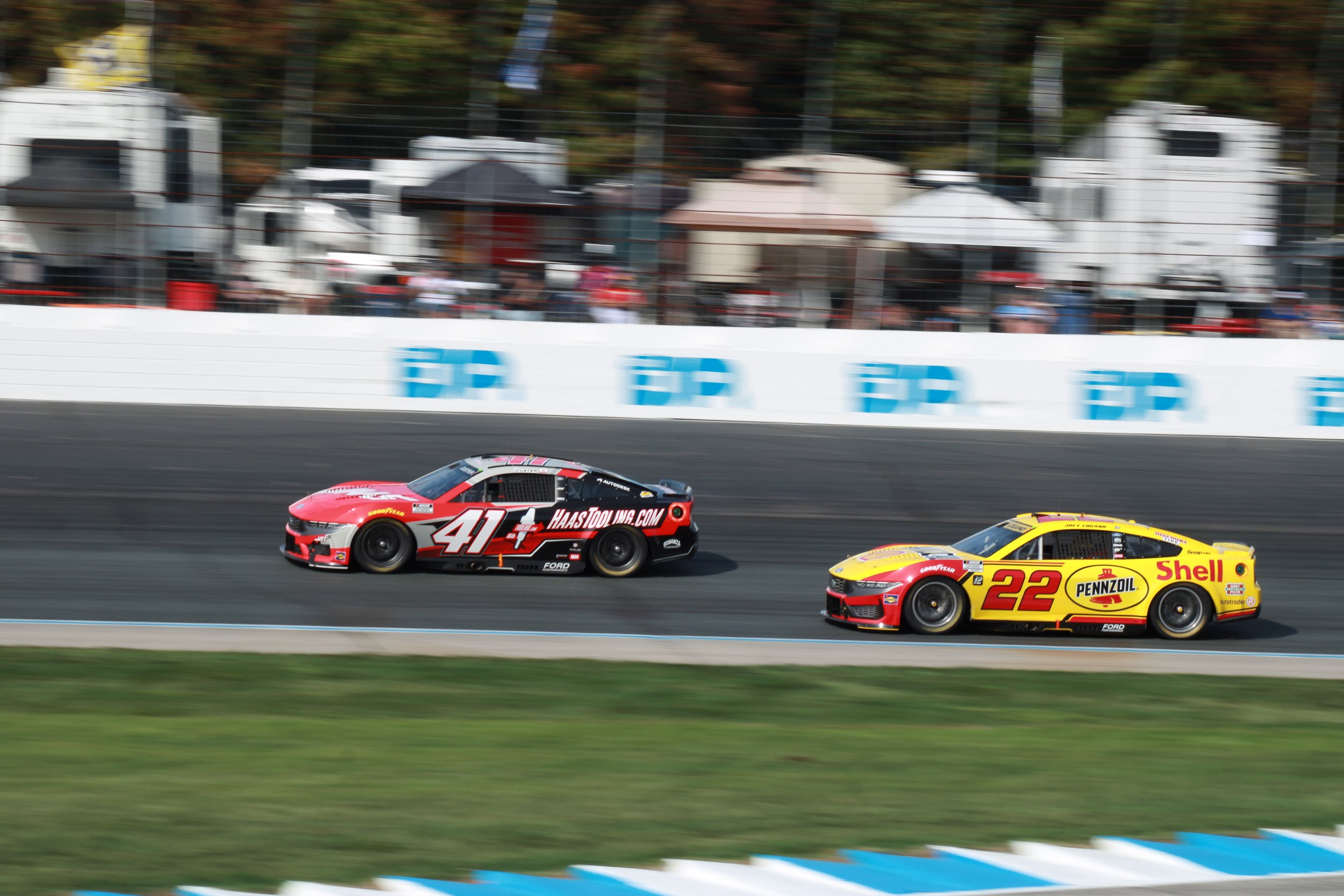 Two racing cars on a track, one red with the number 41 and the other yellow with the number 22, speeding past a blurred background of spectators and tents.