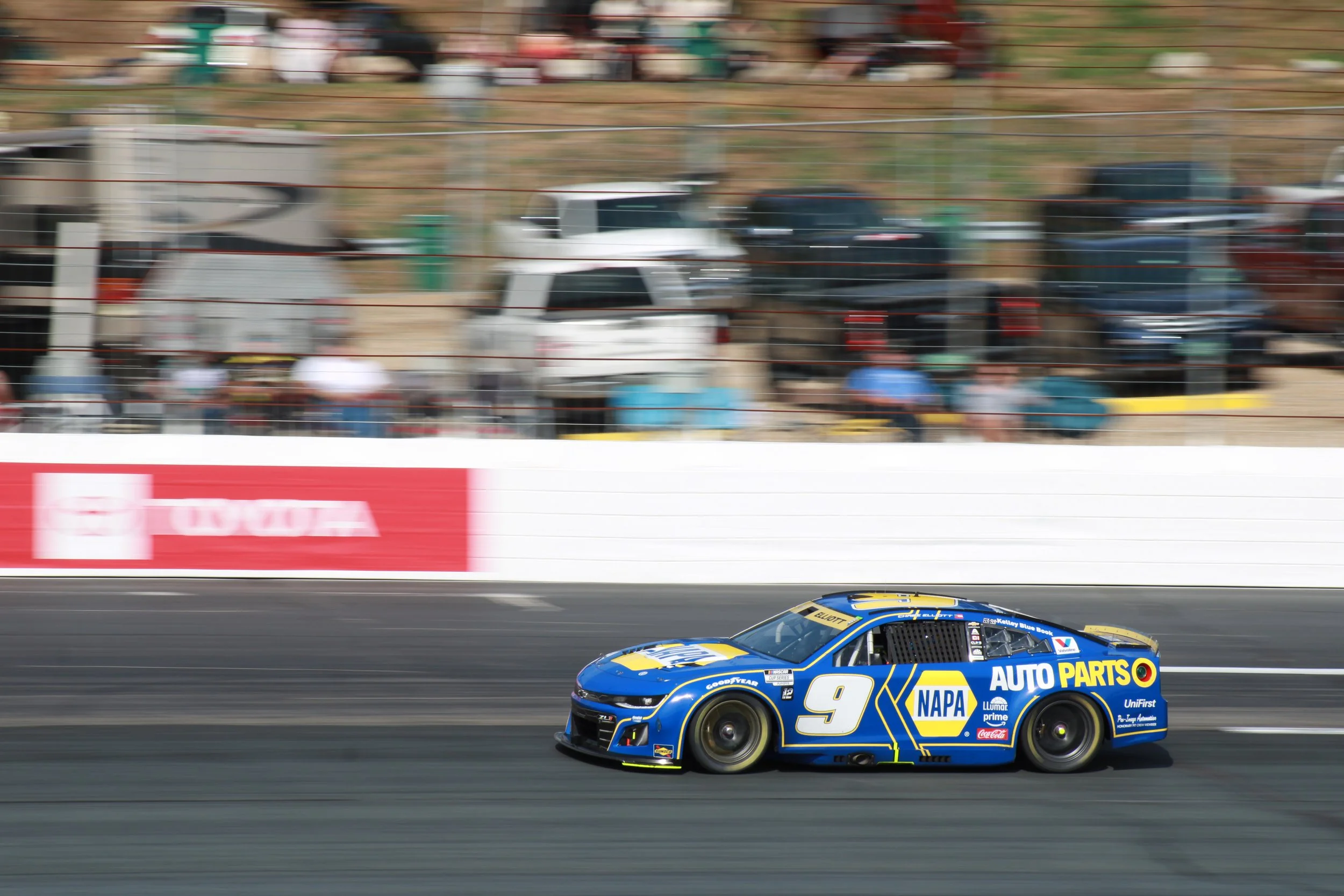 A blue race car with the number 9 and NAPA Auto Parts branding speeding along a racetrack, with a blurred background of parked vehicles and spectators.