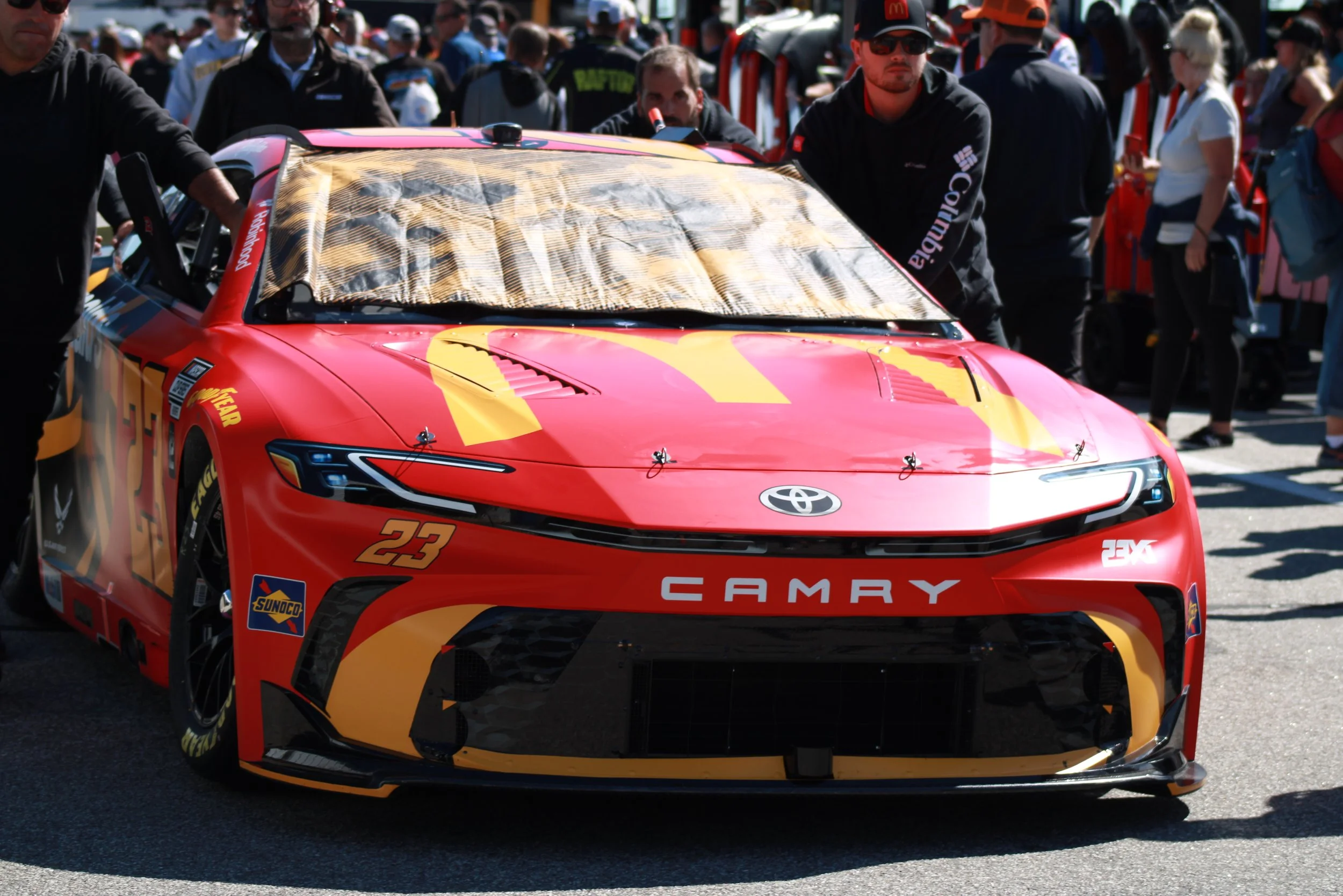 Red Toyota Camry race car with yellow and black accents, number 23, surrounded by a crowd of people at a racing event.